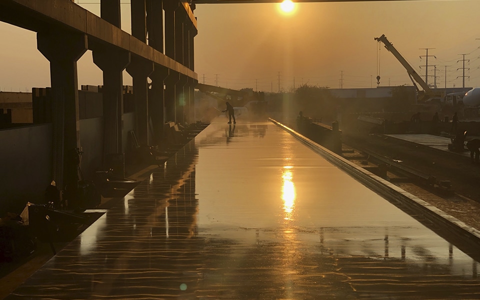 A polished tilting table for precast concrete production reflecting the setting sun, with workers in the background preparing for the next casting cycle in an industrial outdoor setting